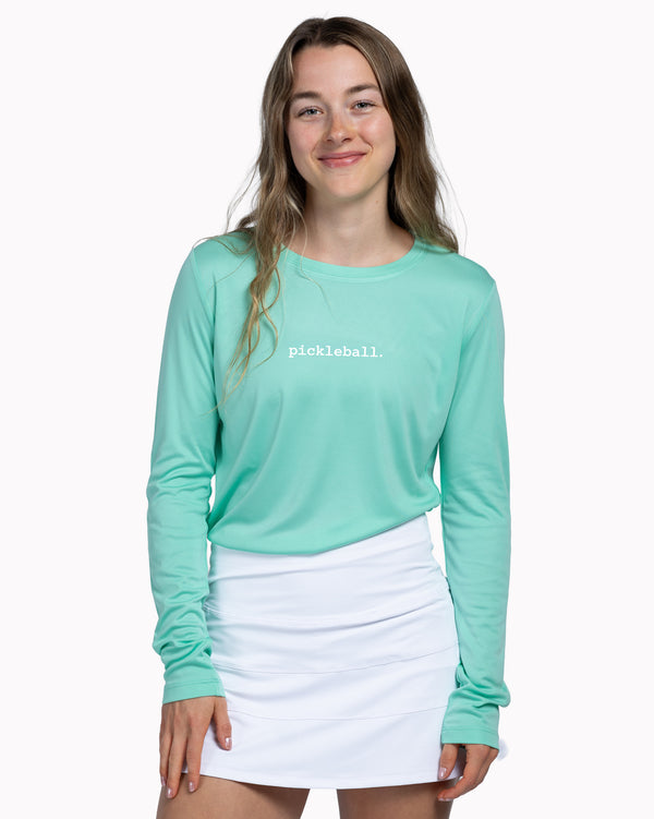 Woman wearing a teal long-sleeve shirt with 'pickleball' text and a white skirt