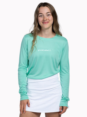 Woman wearing a teal long-sleeve shirt with 'pickleball' text and a white skirt 
