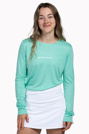 Woman wearing a teal long-sleeve shirt with 'pickleball' text and a white skirt 